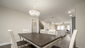 A dining area with a large wooden table and white chairs, a chandelier, and a kitchen in the background. - New to the Vacation Market! Unit 2B Canal Place (Lewes)