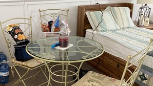 Une petite pièce accueillante, avec un lit, une table en verre et deux chaises. On y trouve des coussins et un panier sur le lit, ainsi qu’une bouteille sur la table.