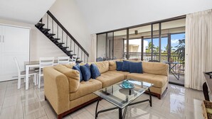 A modern living room with a beige sectional sofa, a glass coffee table, and a staircase leading to a balcony with large windows. - Kona Alii 707 Stunning Top Floor Penthouse w/ Roof Top Deck, AC, & Elevators (KAILUA KONA)