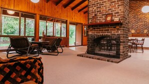 A cozy interior with a stone fireplace, wooden beams, and large windows offering a view of the outdoors. - Woodbine Homestead a Bush and Beach Retreat with a comfortable cosy feel (Hallidays Point)