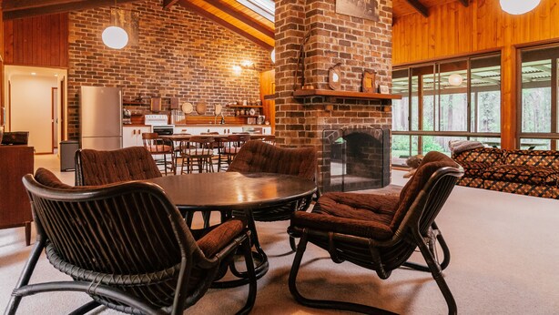 A rustic interior with a brick fireplace, wooden beams, and a dining area with wicker chairs. - Woodbine Homestead a Bush and Beach Retreat with a comfortable cosy feel (Hallidays Point)