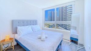 Ein Schlafzimmer mit einem großen Bett, Blick auf hohe Gebäude und einem weißen Nachttisch mit einer Lampe.