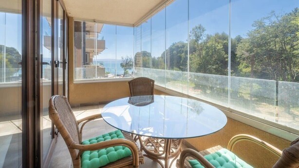 A balcony with a glass table and wicker chairs, overlooking a scenic view.