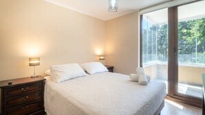 A bedroom with a large bed, two bedside lamps, a nightstand with drawers, and a view of a balcony through a glass door.