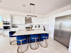 A modern kitchen with white cabinets, a stainless steel refrigerator, a marble island, and blue bar stools.