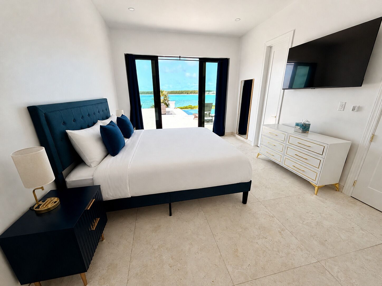 A modern bedroom with a large bed, a dresser, a TV, and a view of the ocean.