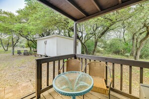 Una terraza techada con sillas de mimbre y una mesa de vidrio, con vista a un área boscosa y un cobertizo pequeño.