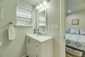 Un baño con un lavabo blanco, un espejo y una ventana con persianas. En la habitación contigua hay una cama con ropa de cama estampada.