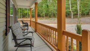 A covered porch with wooden railings, two rocking chairs, and a view of a forested area. - West End Home, Close to Pinehurst Golf Courses! (West End)