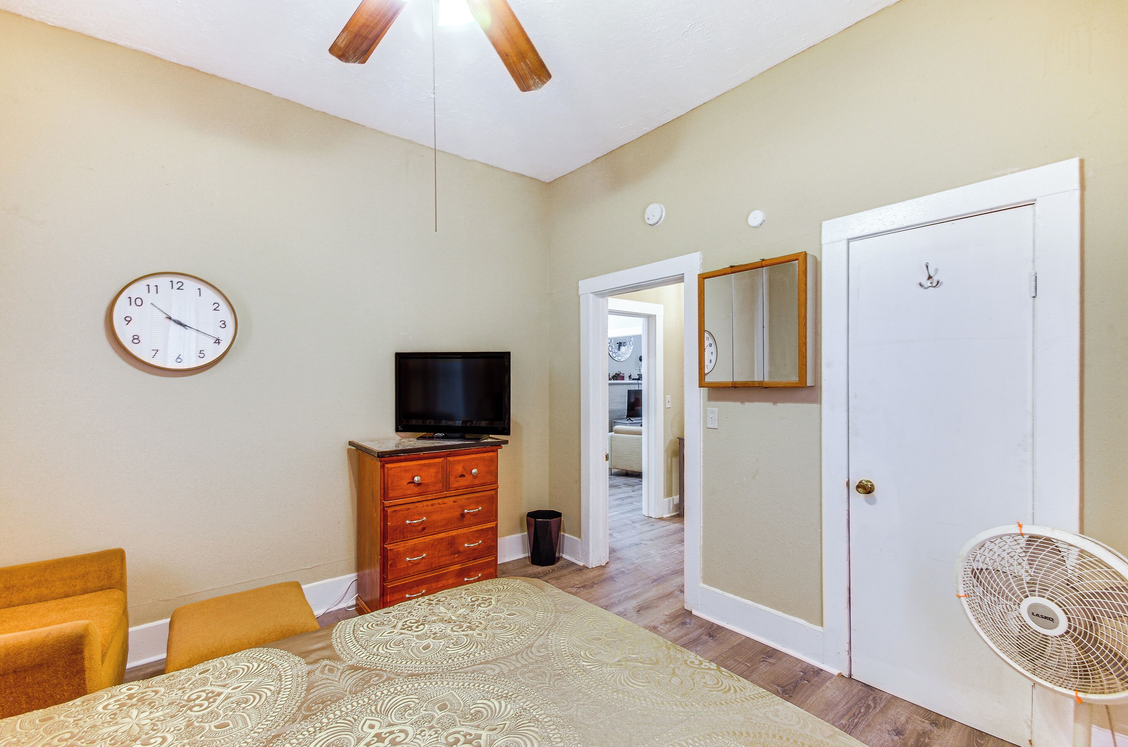 A bedroom with a bed, dresser, TV, clock, and a fan.
