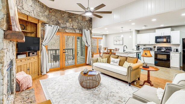 A spacious living area with a stone wall, a ceiling fan, a sofa, a coffee table, and a kitchen in the background. - Golf Course Views: Condo in Blue Ridge Mountains! (Burnsville)