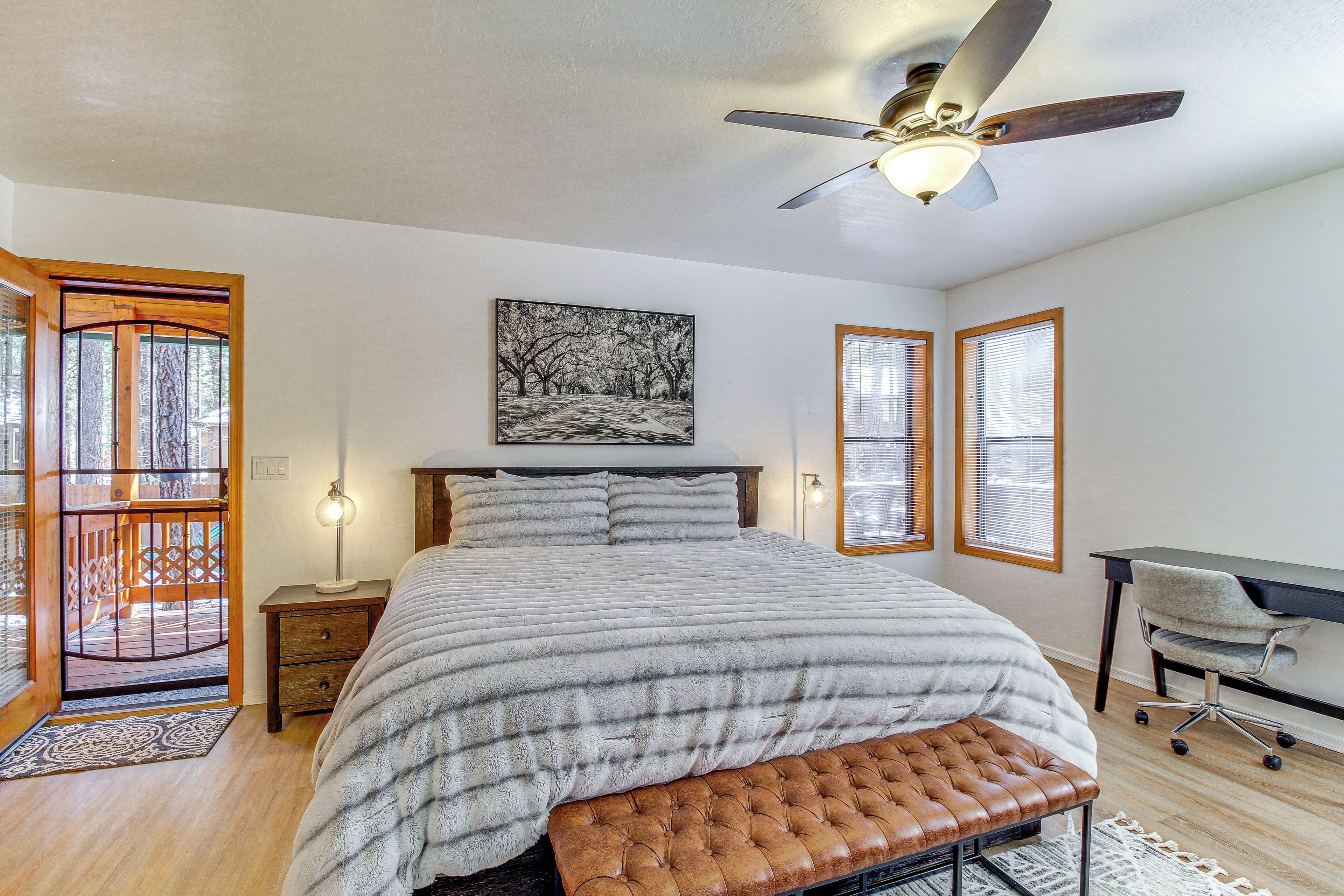 A bedroom with a large bed, a ceiling fan, a desk, and a window with blinds.