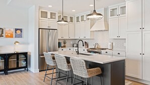 A modern kitchen with white cabinets, a large island with bar stools, stainless steel appliances, pendant lighting, and a backsplash with a geometric pattern. - Persian Bliss 10 Bedroom Townhouse (St. George)