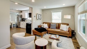A modern living room with a brown leather sofa, a white armchair, and a round coffee table. A kitchen is visible in the background with white cabinets and a refrigerator. - Stylish 'crosby Cottage' - 1 Mi to Serpent Lake! (Crosby)