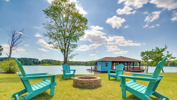 A lakeside deck with turquoise Adirondack chairs, a stone fire pit, and a blue cabin with a dock.