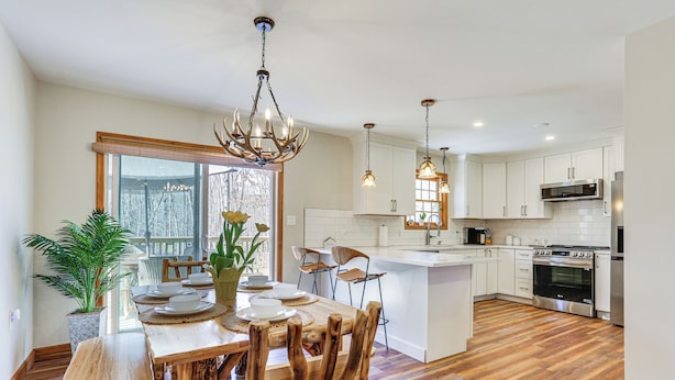 A modern kitchen with wooden flooring, white cabinetry, and a central island. There are pendant lights above the island and a large window providing natural light. - Forest-view Deck & Fire Pit: Poconos Escape! (East Stroudsburg)