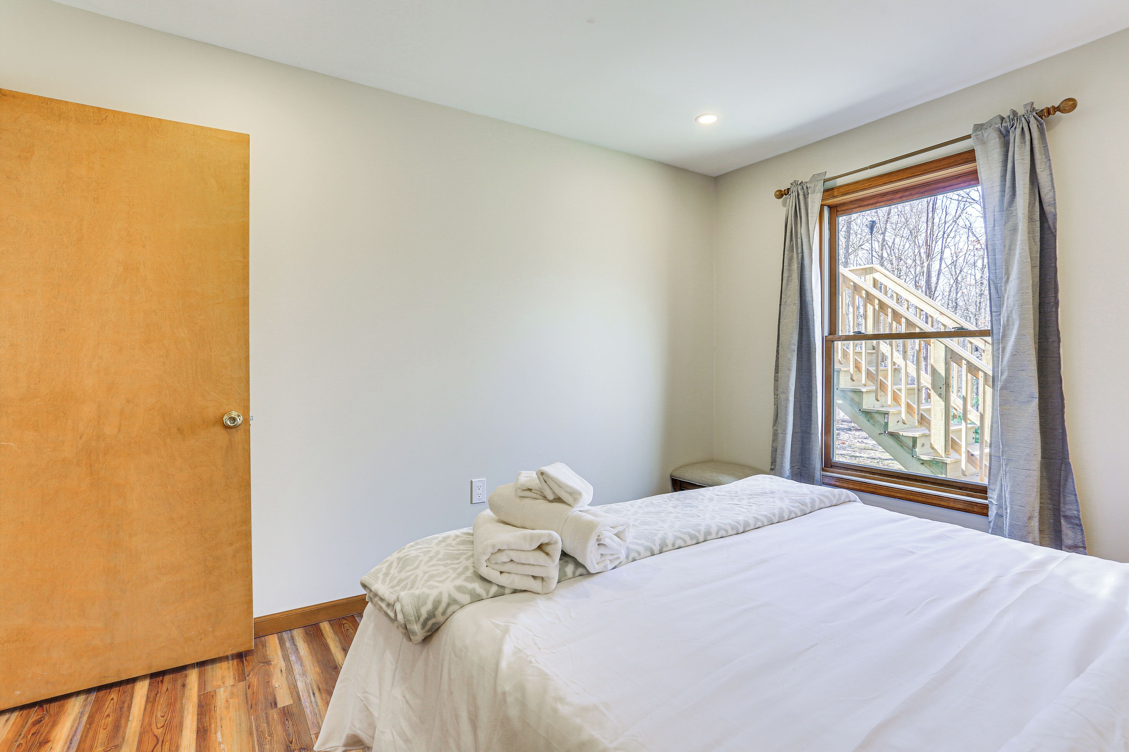 A bedroom with a bed, a wooden door, a window with a view of a wooden staircase, and a small table.