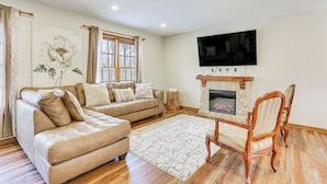 A living room with a beige sectional sofa, a wooden floor, a stone fireplace, and a flat-screen TV mounted above it. - Forest-view Deck & Fire Pit: Poconos Escape! (East Stroudsburg)