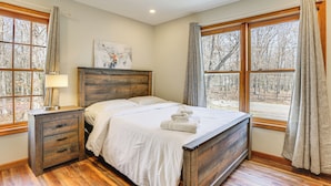A bedroom with a wooden bed, a nightstand, a lamp, and large windows with curtains. - Forest-view Deck & Fire Pit: Poconos Escape! (East Stroudsburg)