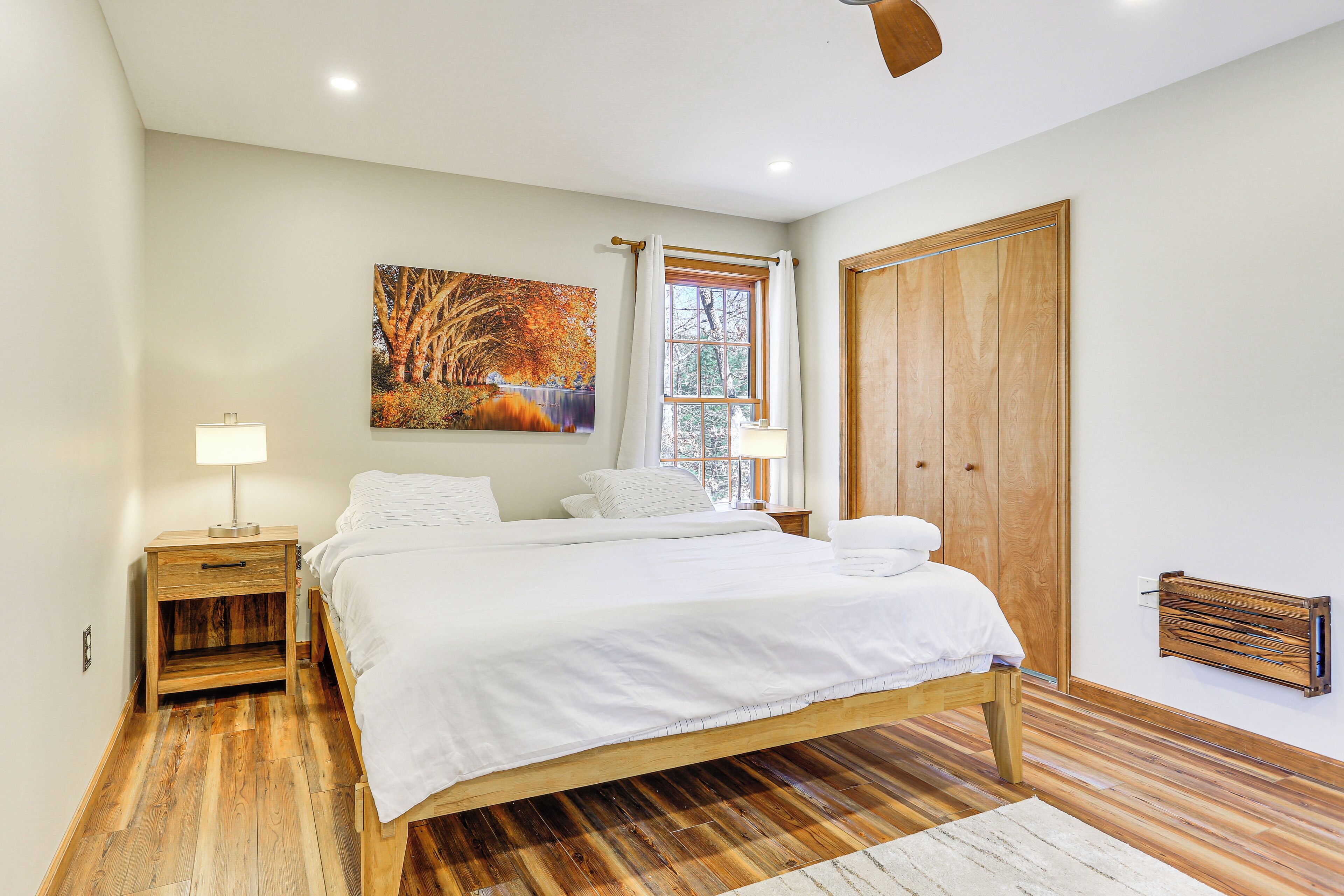 A bedroom with a wooden bed, a nightstand with a lamp, a window with curtains, a wooden wardrobe, and a painting on the wall.