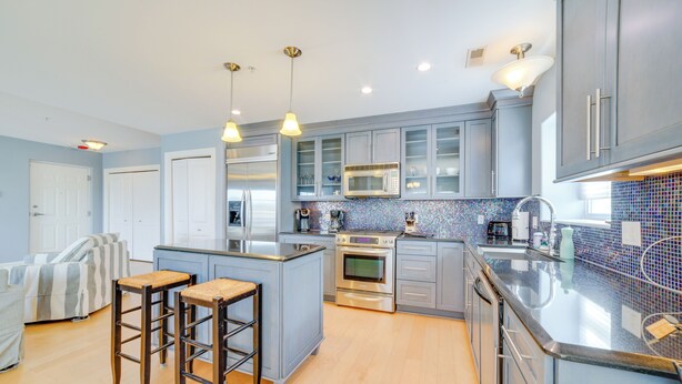 Una cocina moderna con armarios grises, una isla central con taburetes, electrodomésticos de acero inoxidable y un salpicadero de azulejos azules.