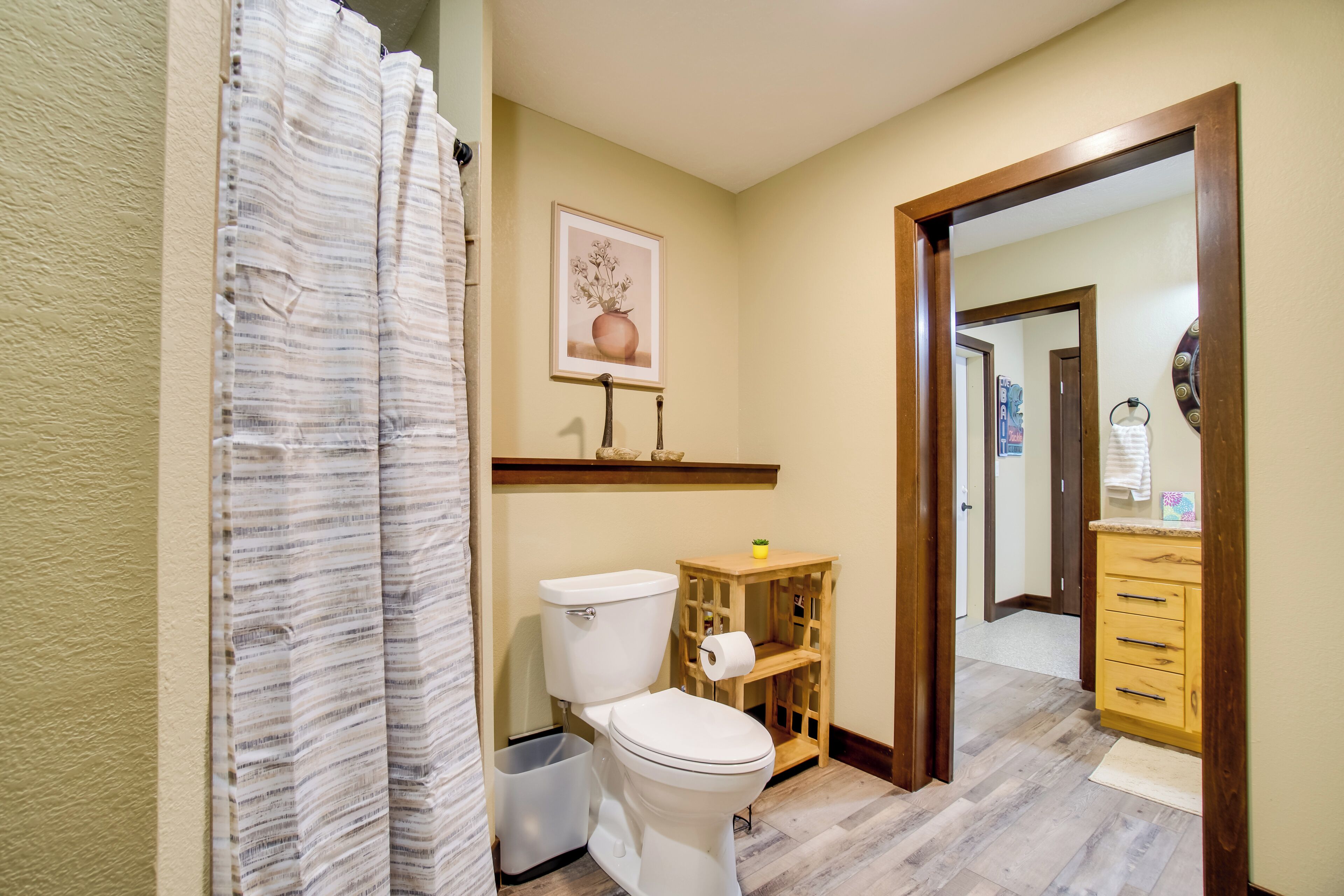 A bathroom with a toilet, a wooden shelf, a trash bin, and a shower curtain.