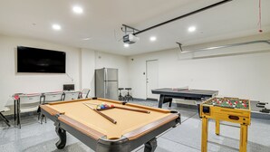 A pool table, foosball table, and a television mounted on the wall in a room with white walls and a ceiling projector.