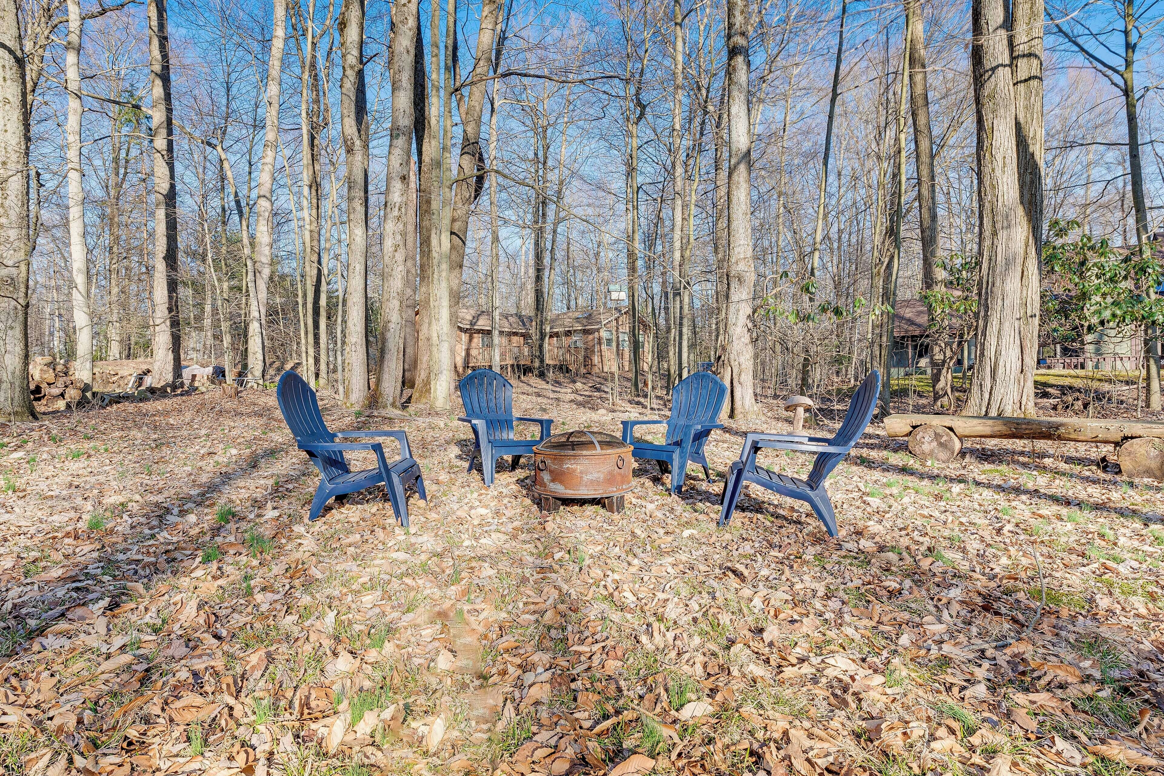 A forested area with a fire pit and three blue chairs arranged around it.
