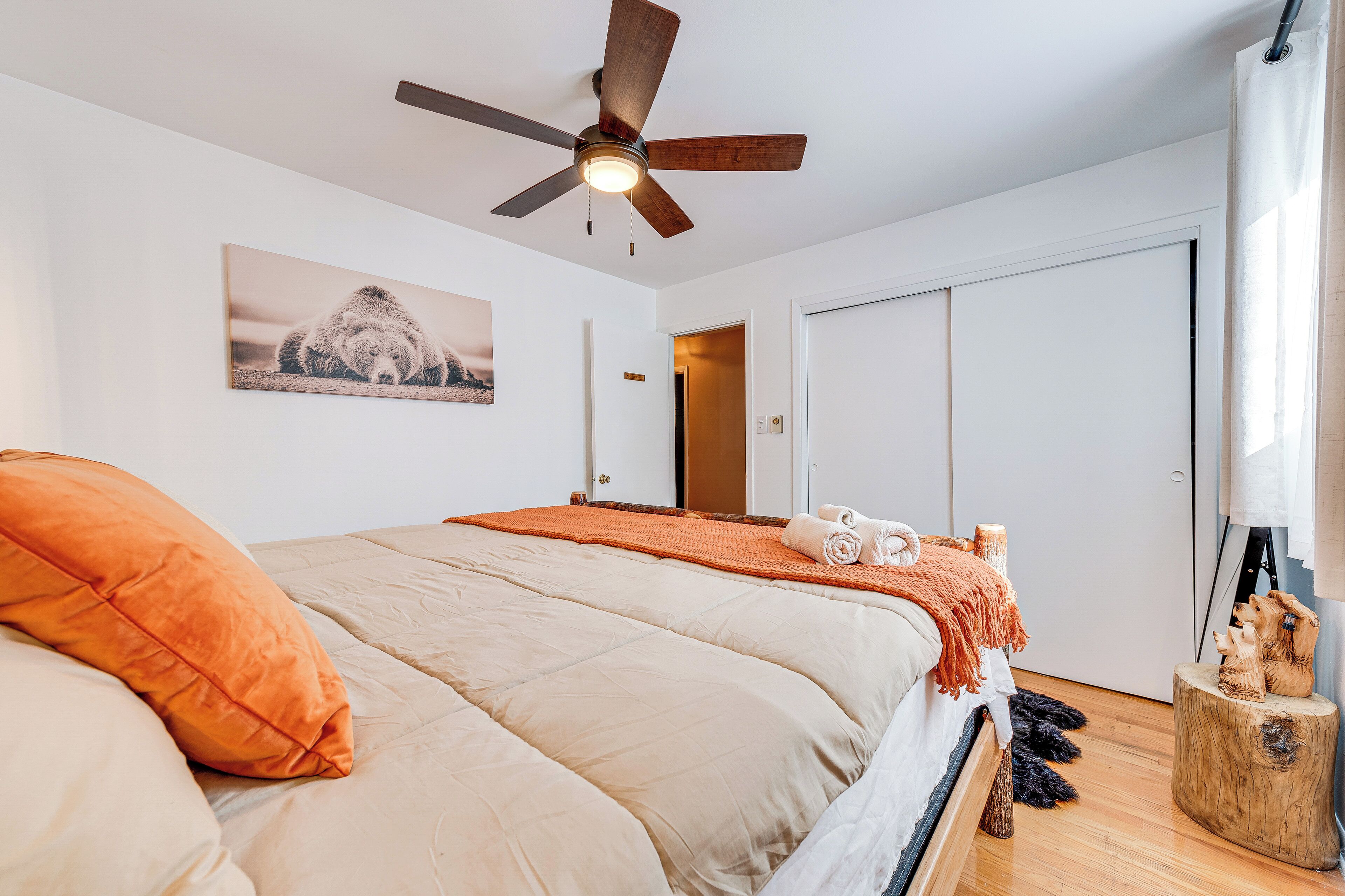Ein Schlafzimmer mit Bett, Deckenventilator, Holz-Kopfteil, einem Gemälde, einer Tür und einem Fenster mit Vorhängen.