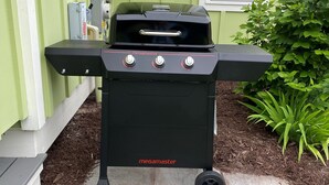 Musta Megamaster-grilli, jossa on kolme säätönuppia, on sijoitettu laatoitetulle terassille, jonka pohjalla on kuorikate. Grillin edessä on vihreä kasvihuone, jossa on ikkuna ja sälekaihtimet.