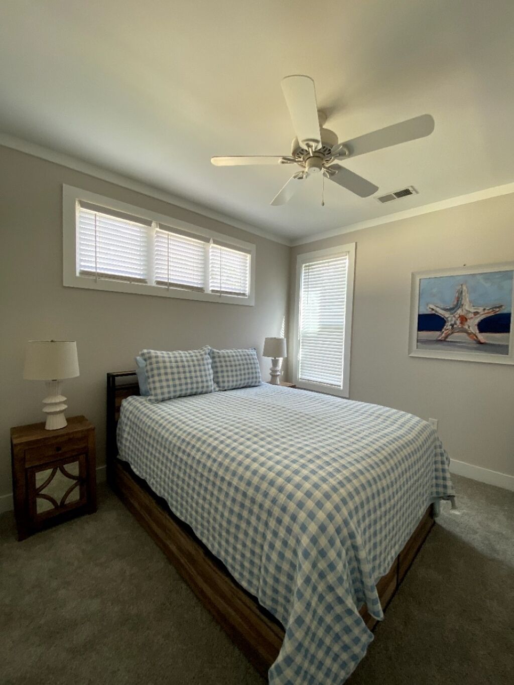 A bedroom with a bed, a ceiling fan, two windows with blinds, a nightstand with a lamp, and a framed picture on the wall.