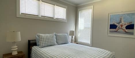A bedroom with a bed, a ceiling fan, two windows with blinds, a nightstand with a lamp, and a framed picture on the wall.