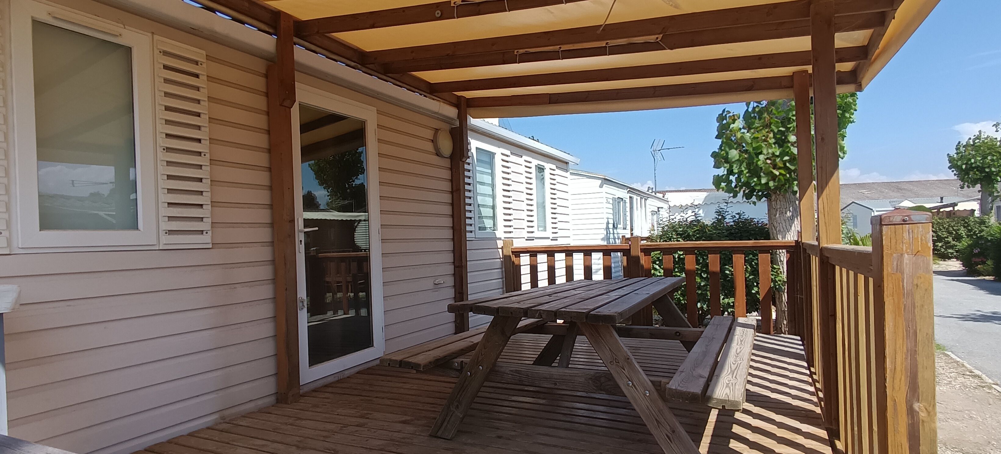 Eine Holzterrasse mit Picknicktisch und überdachtem Bereich, angrenzend an ein Haus mit einer Schiebeglastür.