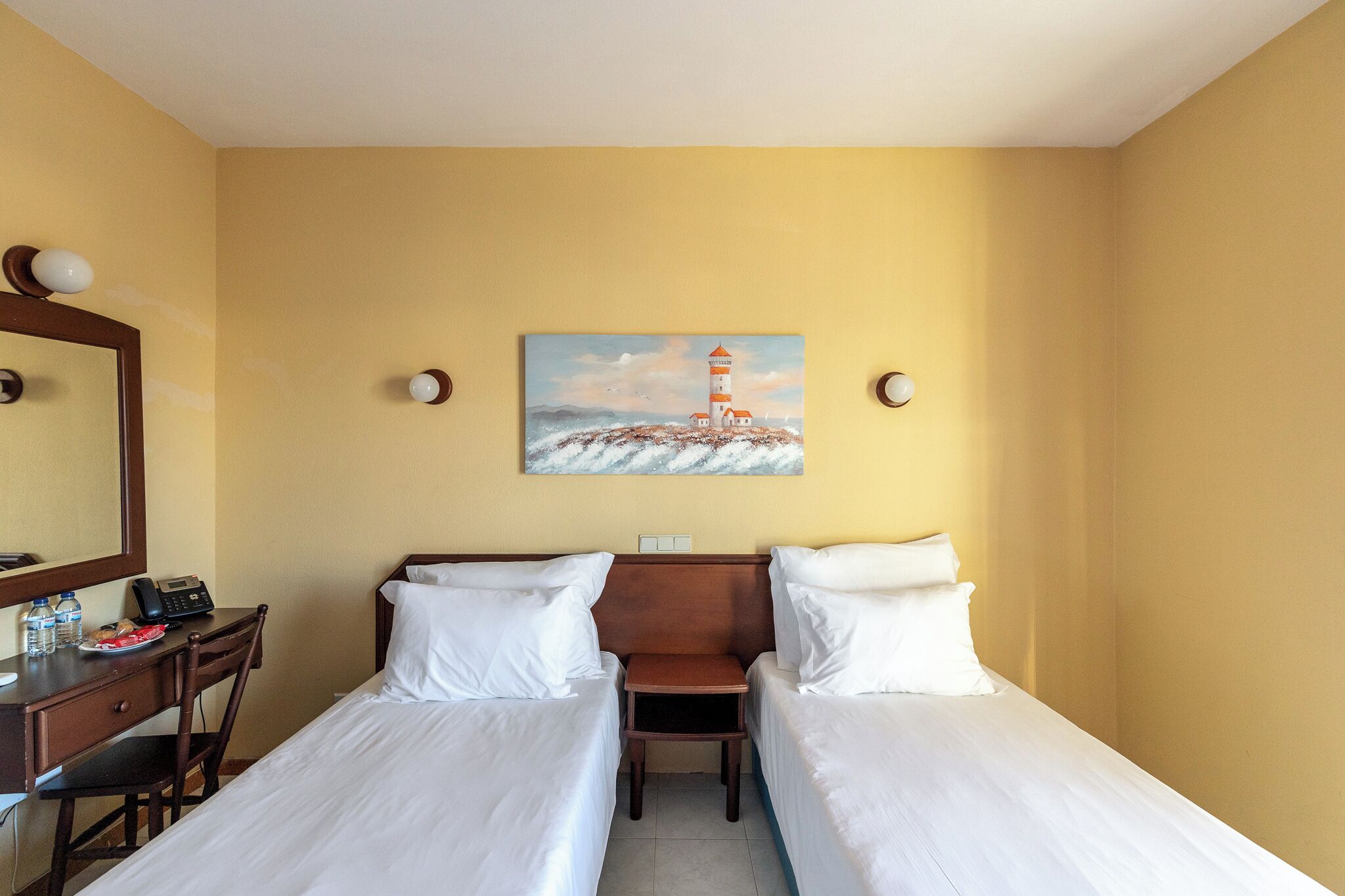 Ett hotellrum med två separata sängar, ett skrivbord med en stol, en telefon och en tavla med ett motiv av en fyr på väggen.