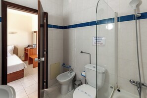 A hotel bathroom with a shower, bidet, toilet, and a bed in the adjacent room.