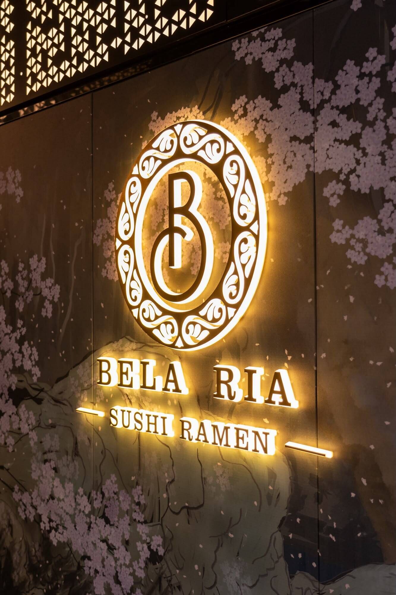 A restaurant sign with a stylized 'B' logo and the name 'Bela Ria Sushi Ramen' illuminated against a backdrop of cherry blossoms.