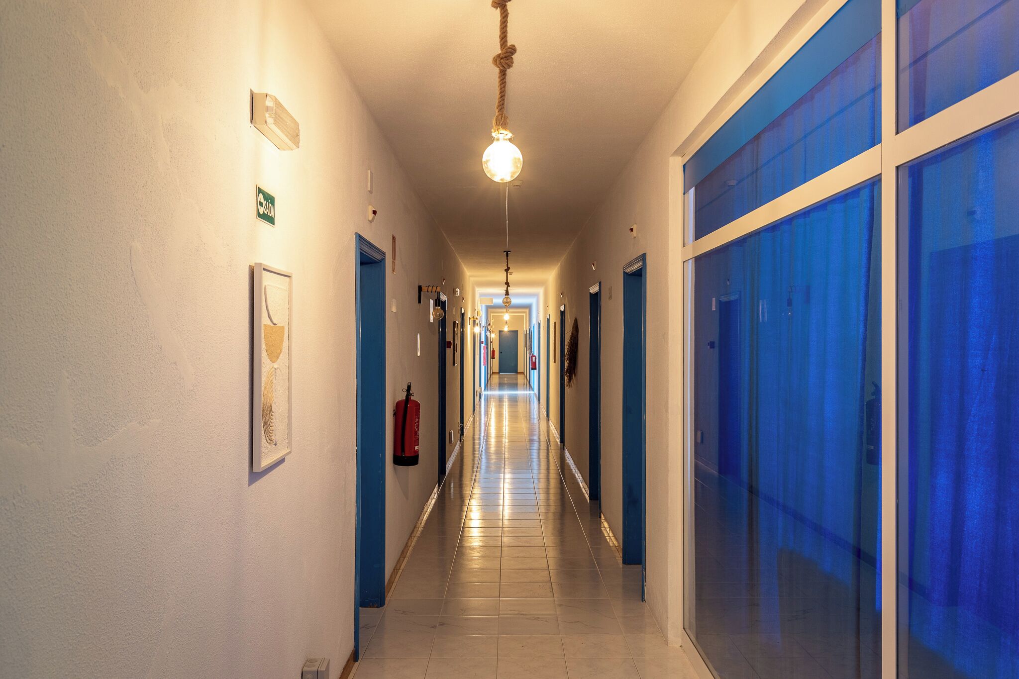 A long, narrow hotel corridor with blue-framed doors, a fire extinguisher, and a hanging light fixture.