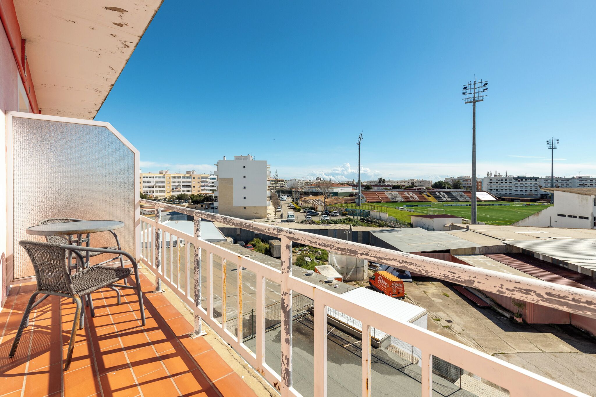 A balcony with a table and chairs, overlooking a sports field and buildings.
