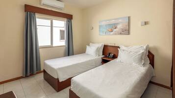 A hotel room with two single beds, a window with curtains, an air conditioner, a painting of boats on the wall, and a bedside table with a phone.