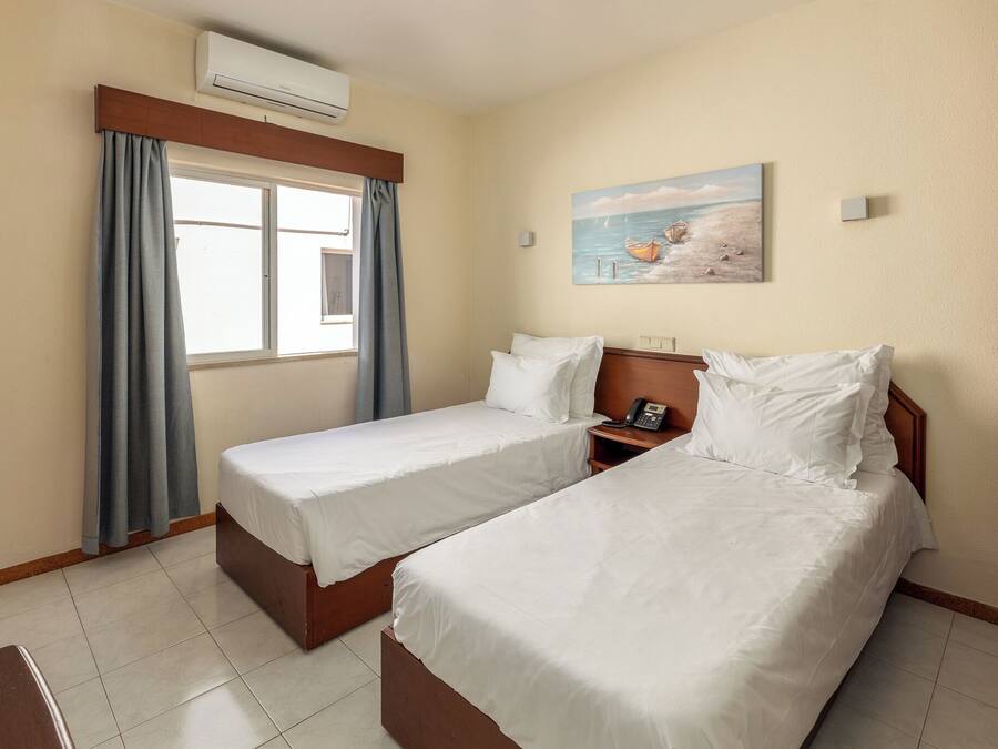 A hotel room with two single beds, a window with curtains, an air conditioner, a painting of boats on the wall, and a bedside table with a phone.