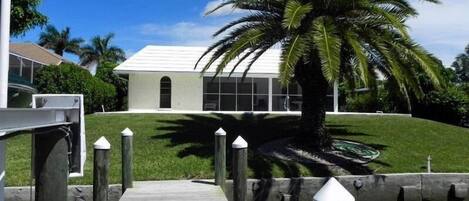 A modern building with a white facade, a wooden dock leading to a water body, and a palm tree in the foreground.