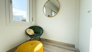 Ein Zimmer mit grünem Sessel, einem gelben Hocker, einem runden Spiegel und einem Fenster mit Blick auf Gebäude.