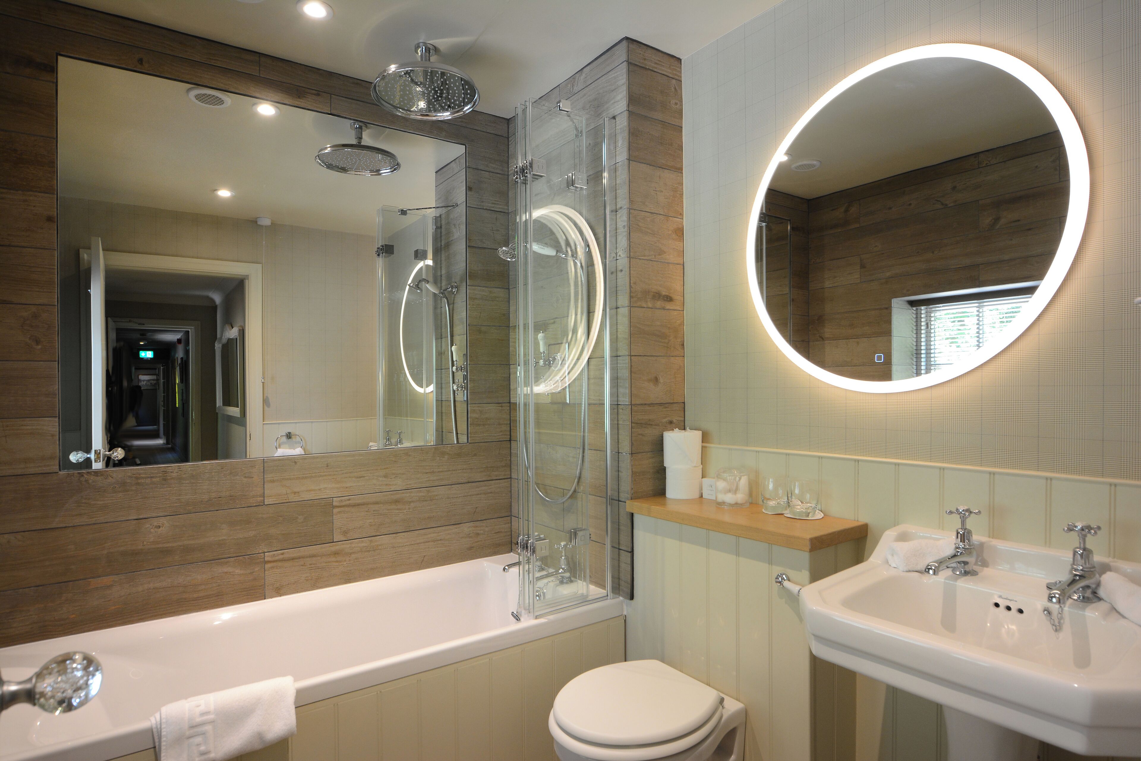 A modern bathroom with a glass-enclosed shower, a large mirror with a circular light, a bathtub, and a sink.