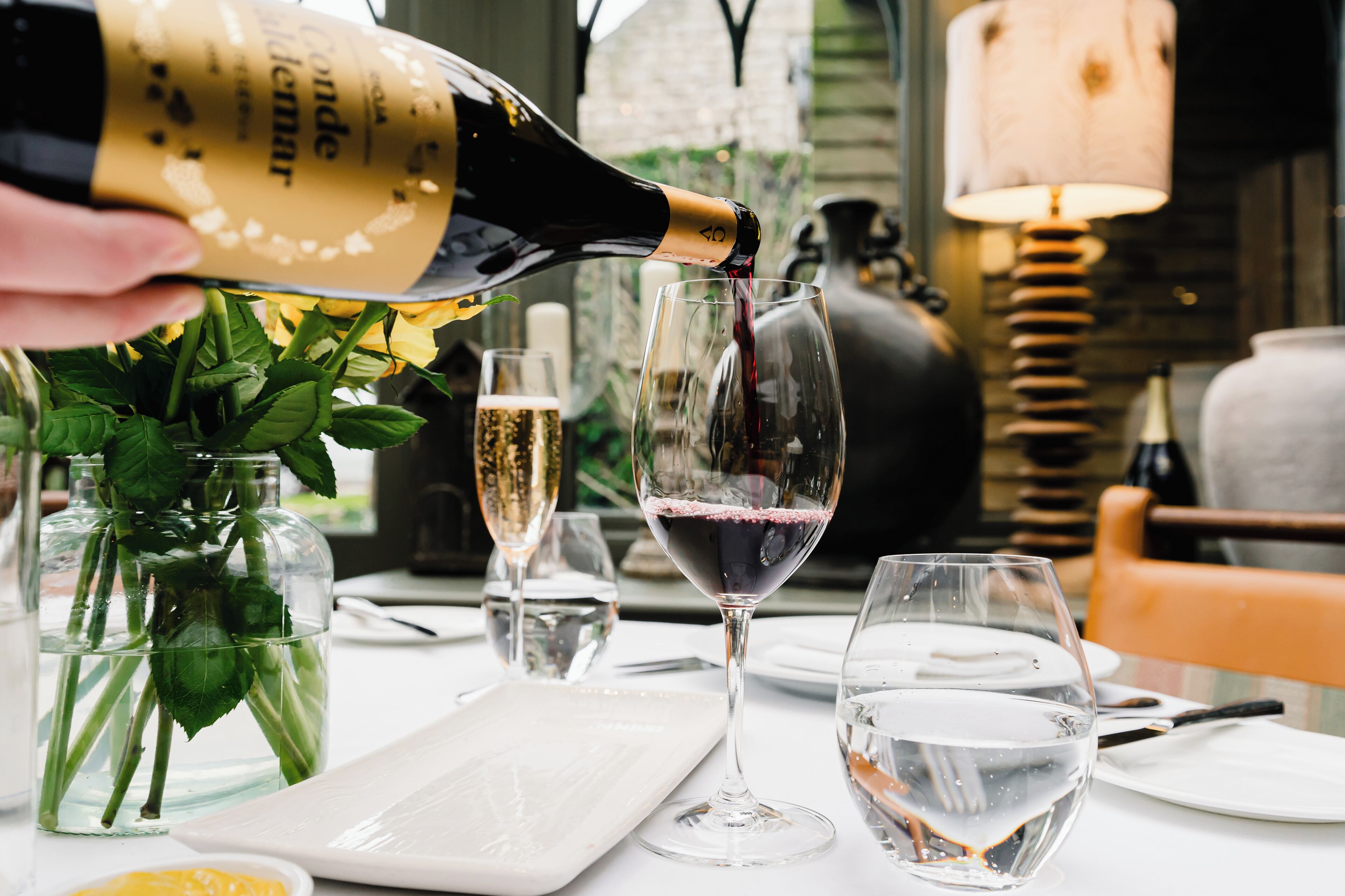 A bottle of champagne being poured into a wine glass, with a vase of flowers and a candle in the background.