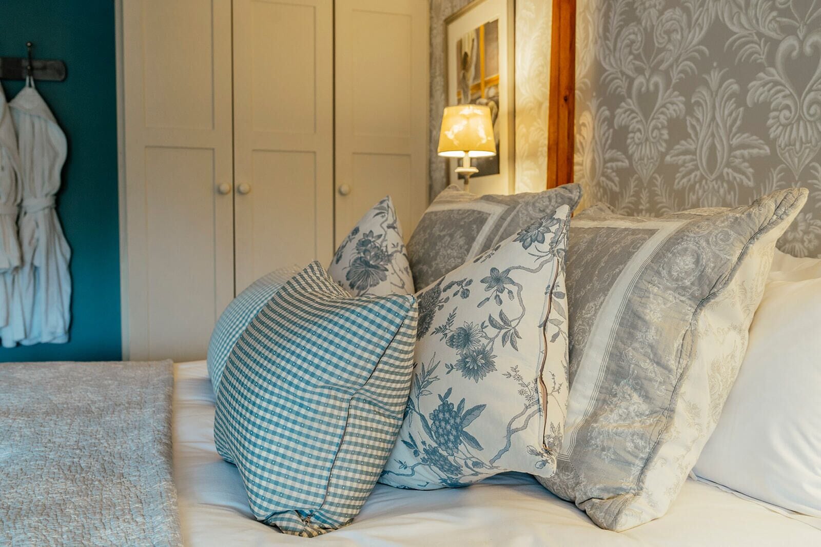 A bed with three pillows, a lamp, and a wardrobe in a room with patterned wallpaper.