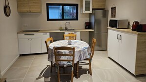 A compact kitchen with a dining table, chairs, a refrigerator, a microwave, and a window with a view of trees. - 2 Pièces, Jardinet Sud, Calme, 1000 m Morgat (Crozon)