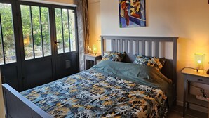 A bedroom with a bed, a painting, a desk with a lamp, and a view through a window. - 2 Pièces, Jardinet Sud, Calme, 1000 m Morgat (Crozon)