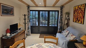A cozy living room with a wooden ceiling, a large window with curtains, a sofa, a table with a nautical-themed tablecloth, and a television on a wooden desk. - 2 Pièces, Jardinet Sud, Calme, 1000 m Morgat (Crozon)