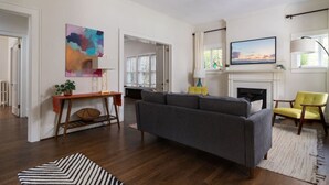 Een ruime woonkamer met een grijze bank, een gele fauteuil en een open haard. Er hangt een schilderij aan de muur en er is een raam met gordijnen.