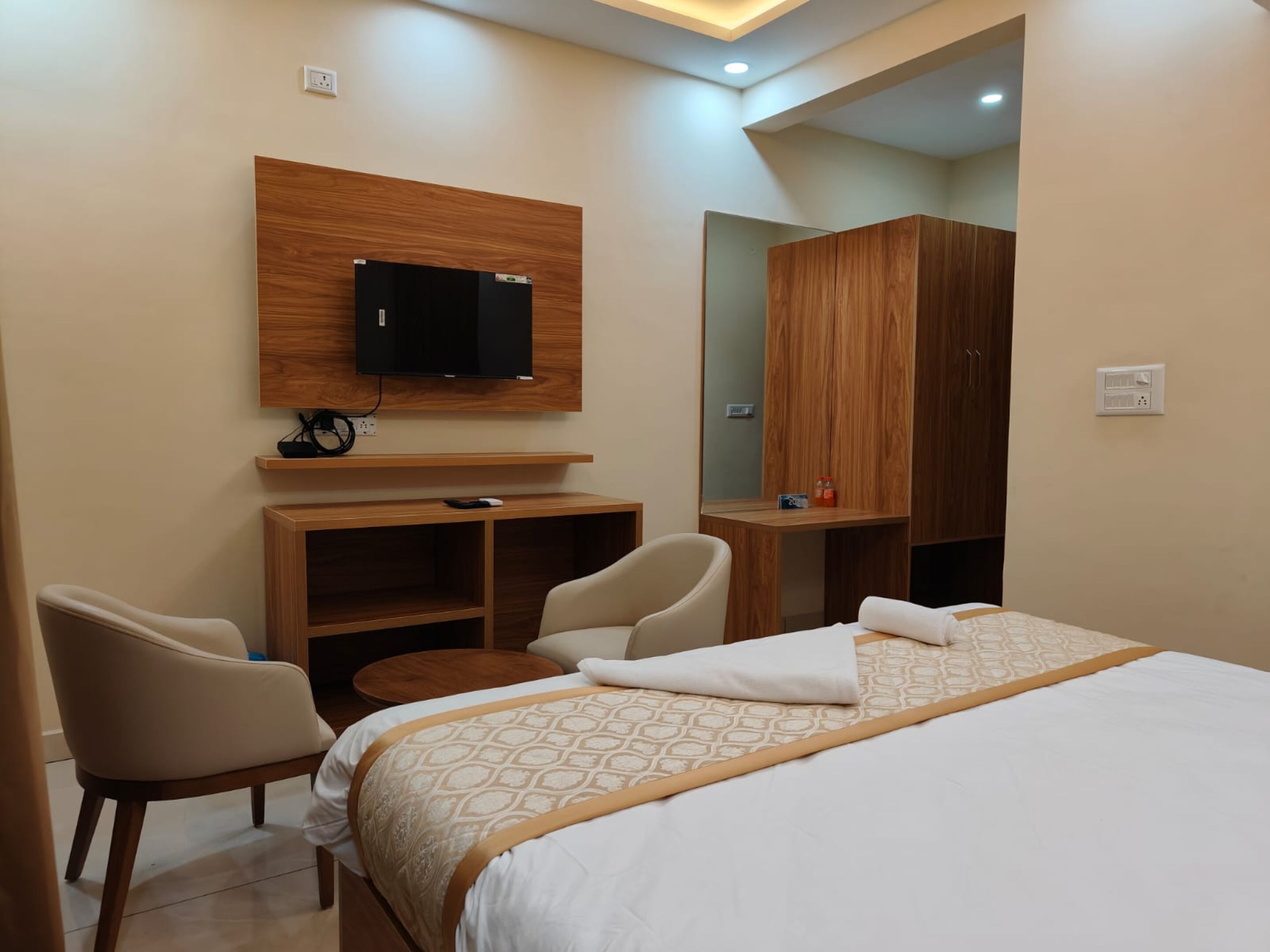 A hotel room with a bed, a desk, a chair, a TV mounted on a wooden panel, and a wardrobe.